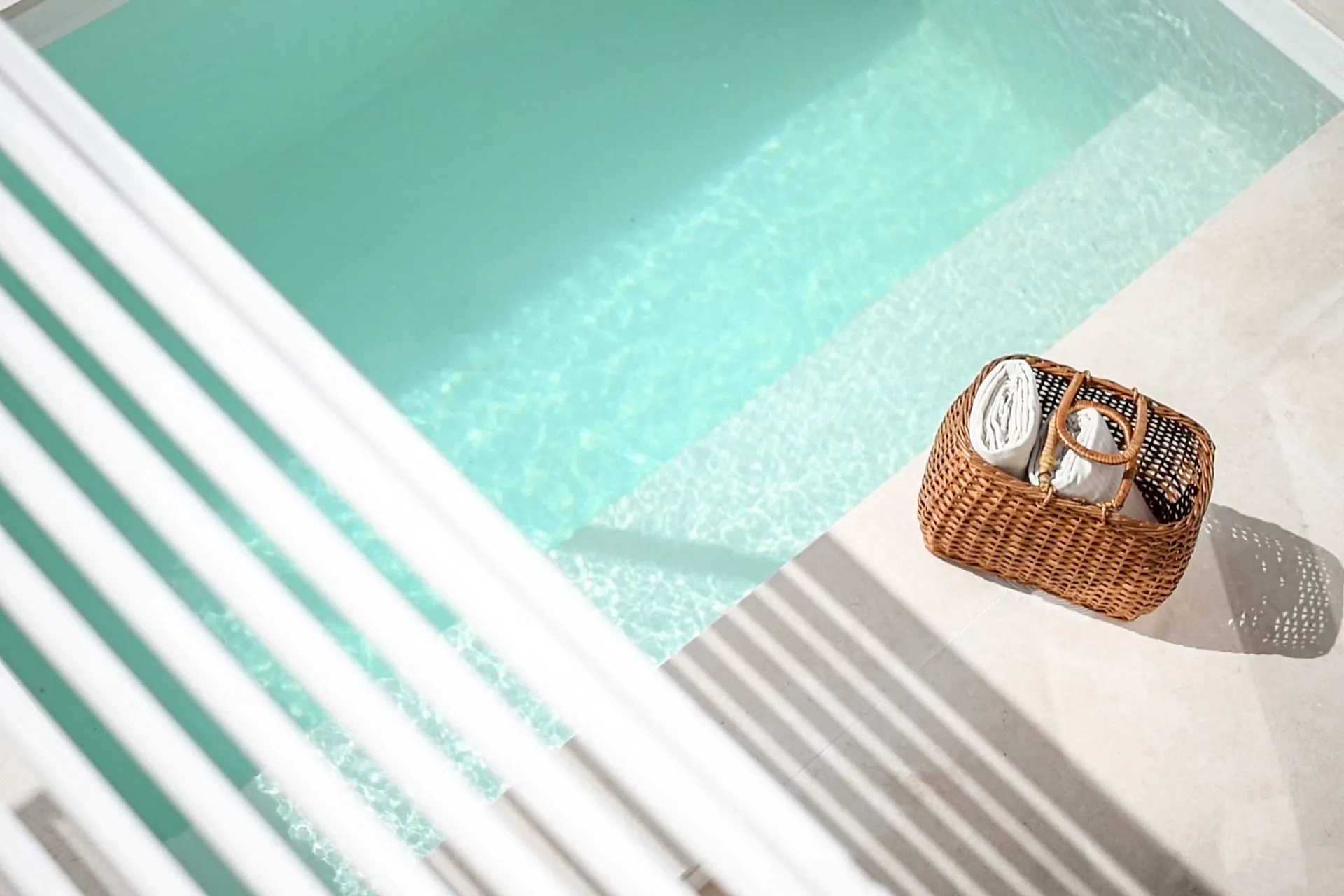 A wicker cane basket filled with rolled-up white towels placed beside a mint green swimming pool.