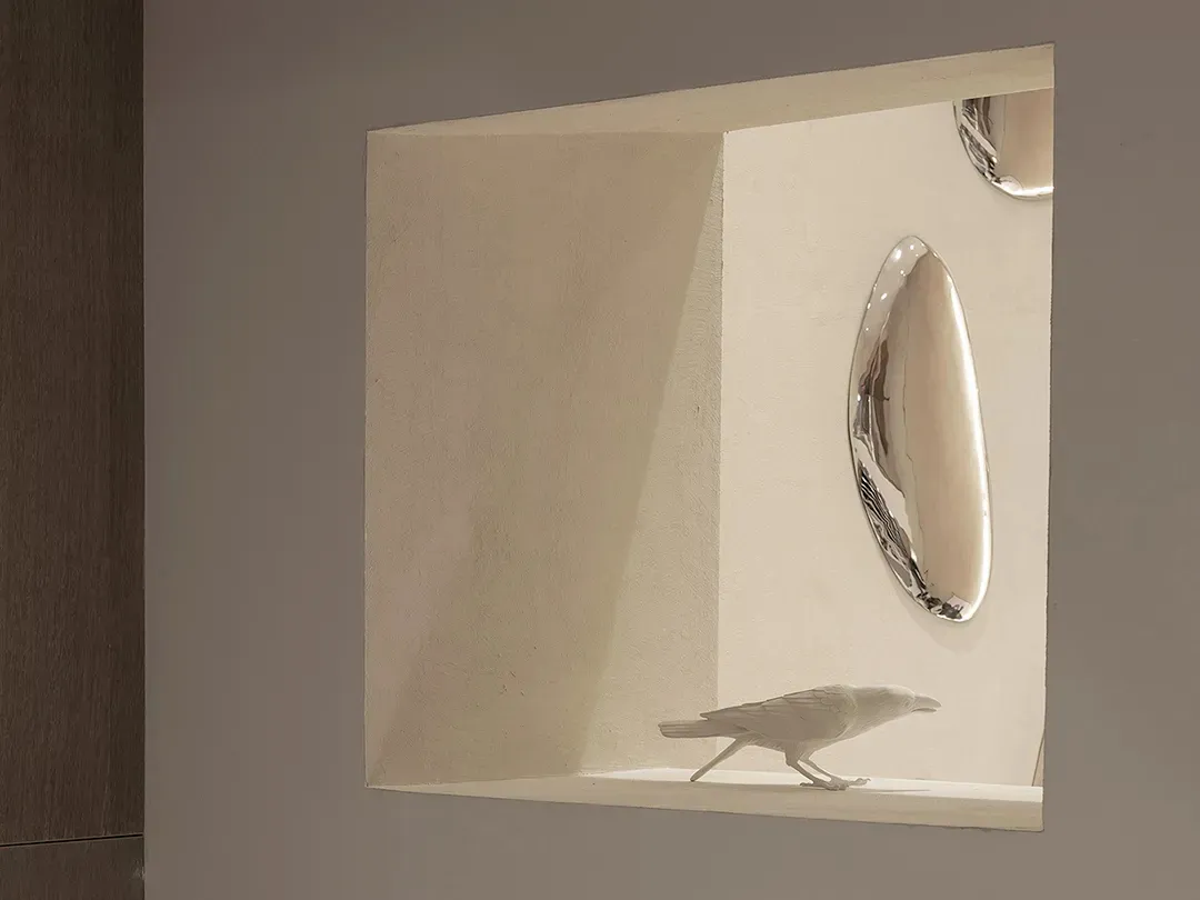 A small ceramic bird figurine sitting in a recessed shelf with reflective metallic plates nearby.