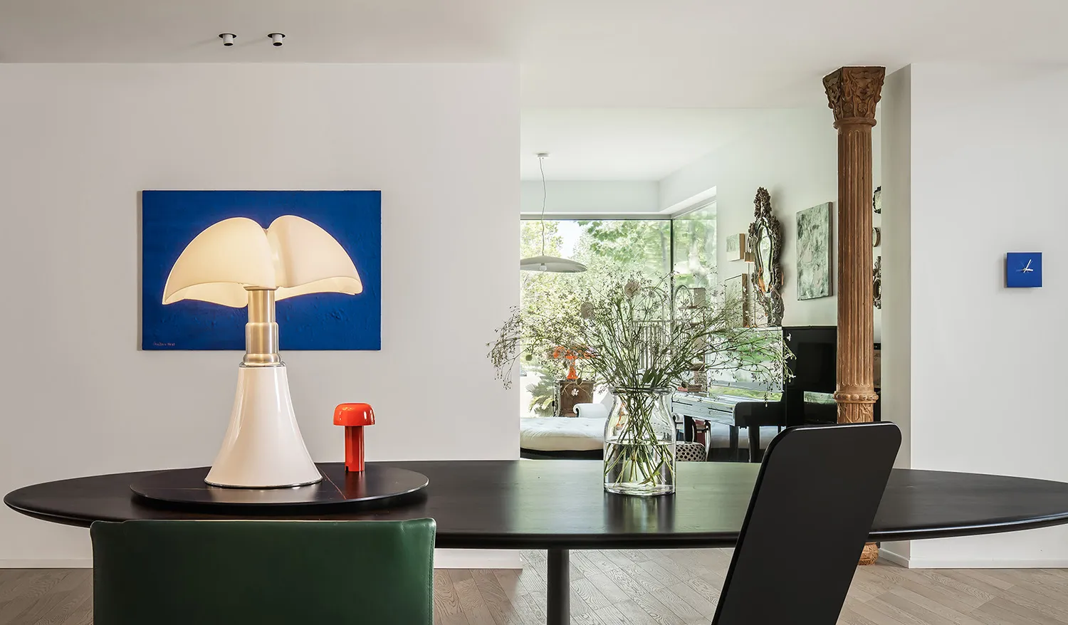 A modern dining room featuring a black oval table, white table lamp, and glass vase with dried flowers.