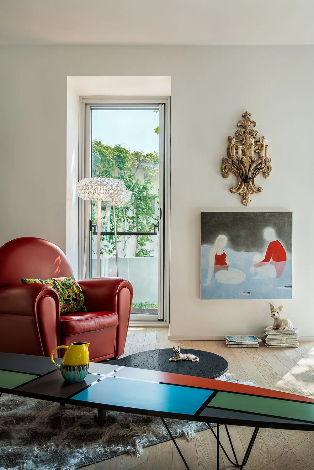 A modern living room featuring a red armchair with a colorful pillow, a colorful coffee table, a decorative sconce, and a pai