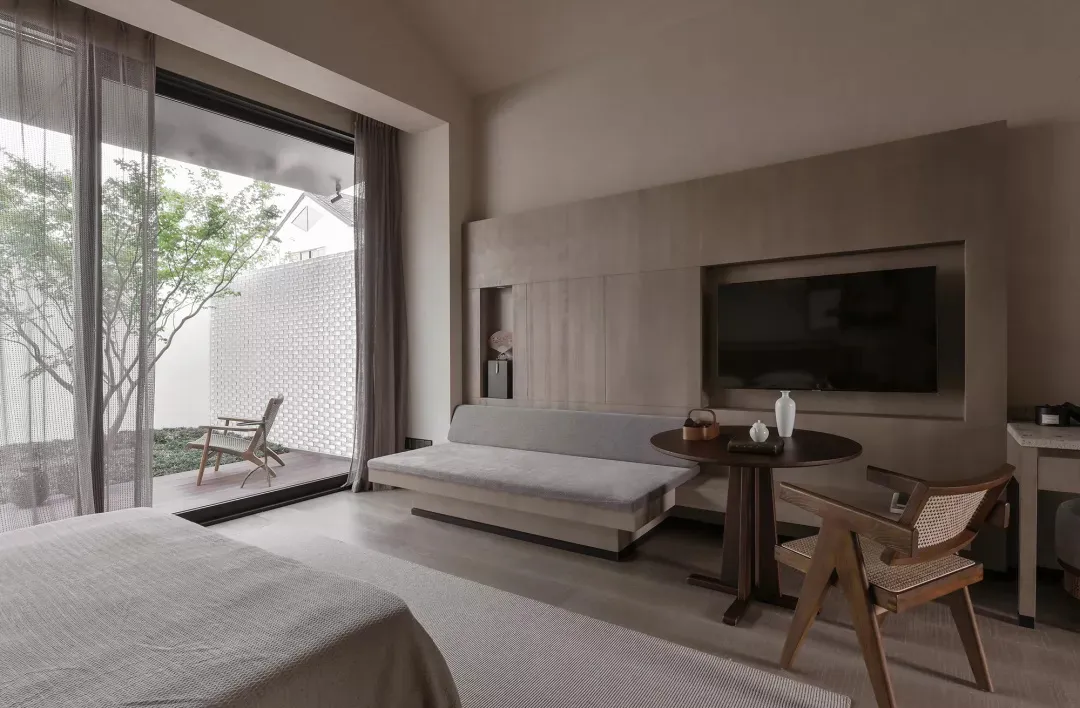 A minimalist bedroom with neutral-toned furniture and a large window overlooking greenery.