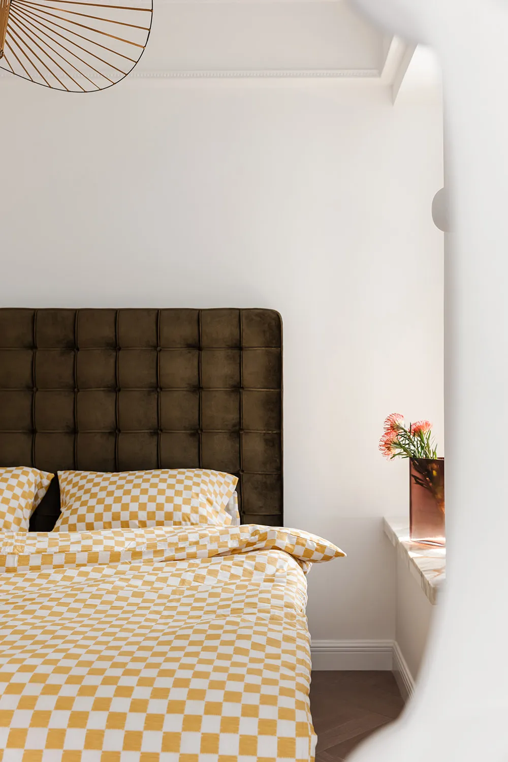 A modern bedroom featuring a velvet upholstered headboard, checkered bedding, and a vase of flowers on a windowsill.
