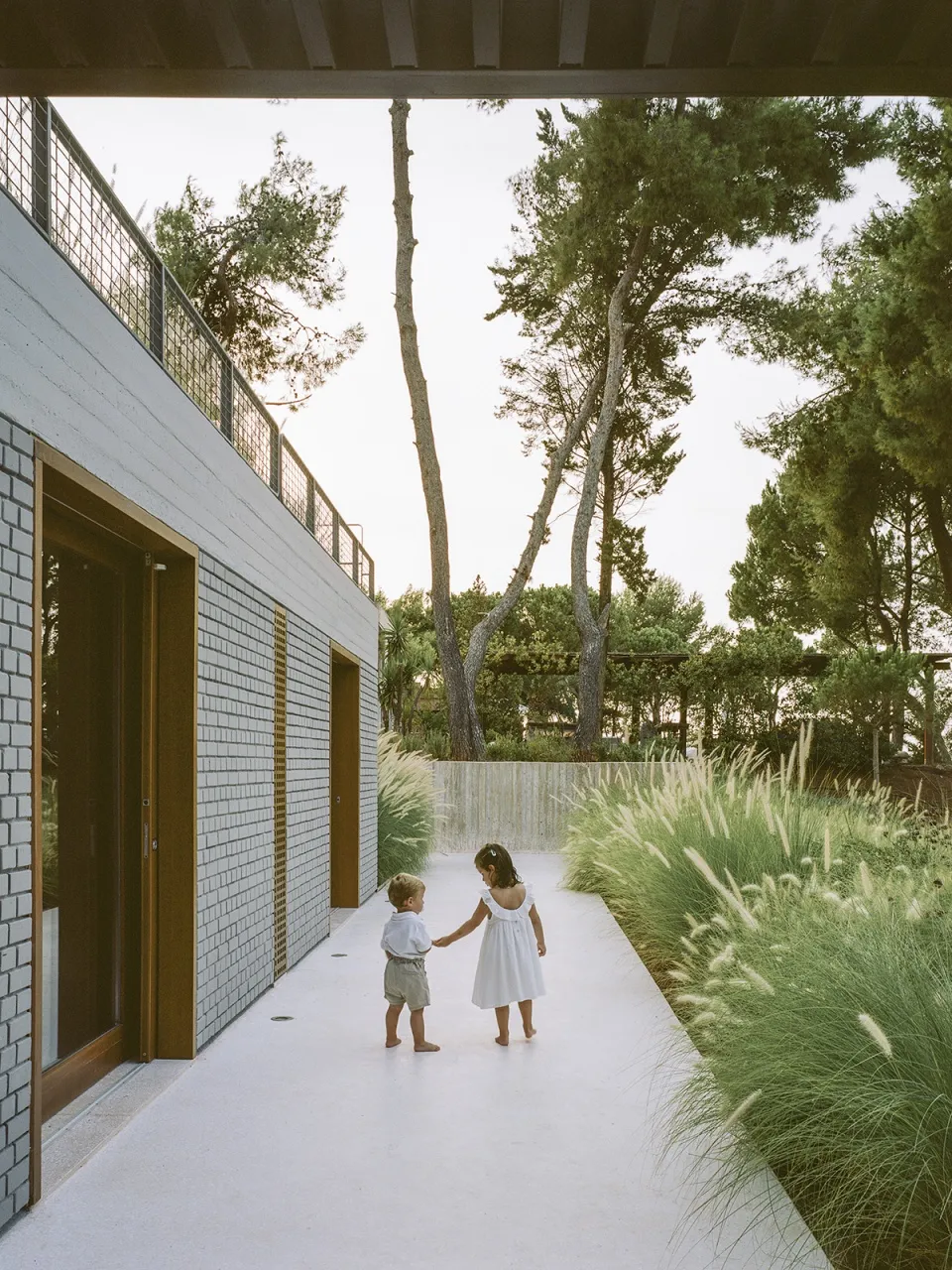 Two children holding hands while walking on a white walkway in a modern concrete building surrounded by greenery.