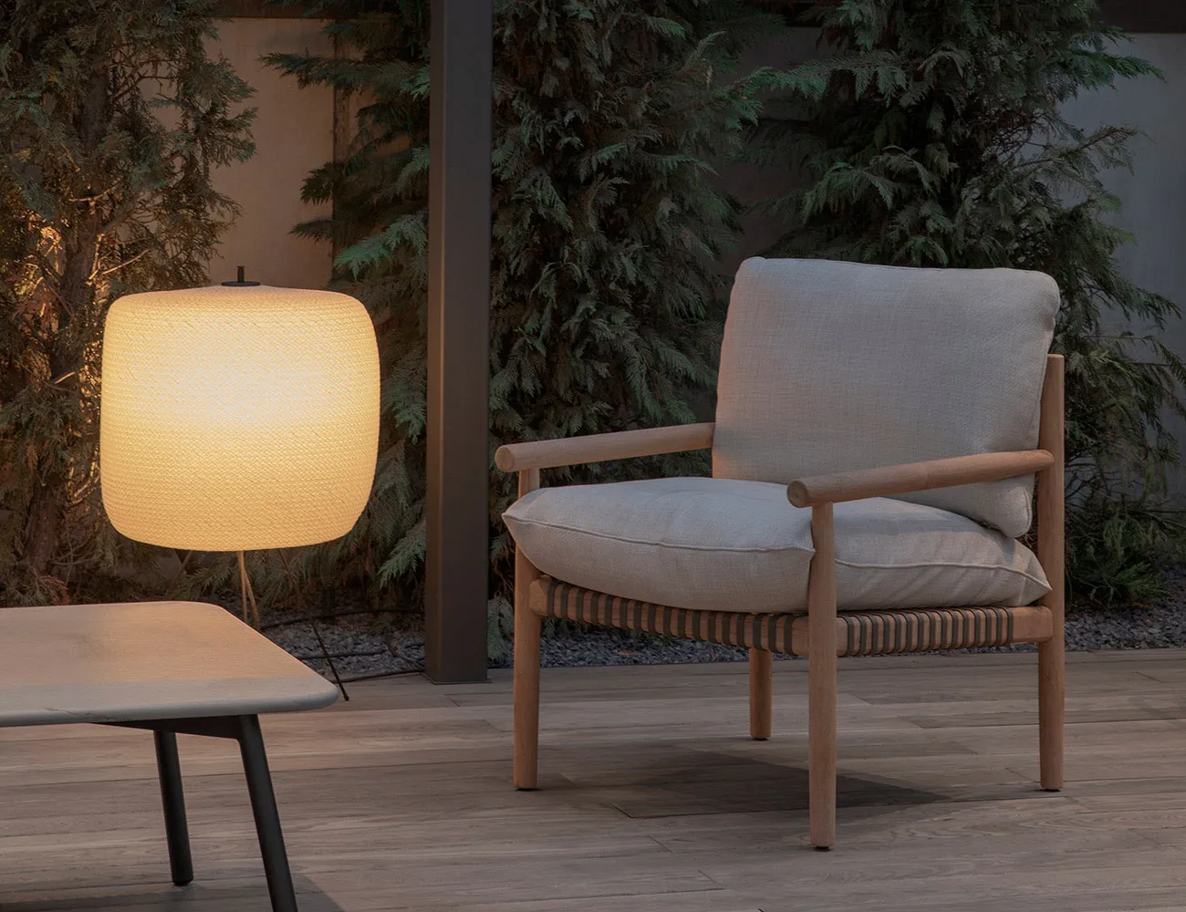 A minimalist outdoor furniture setup featuring a wooden chair with woven armrests and cushions, accompanied by a round, illum