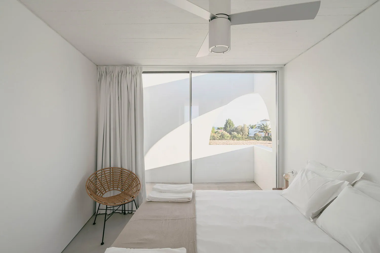 A minimalist white bedroom with a large window showing a view of a garden with palm trees.