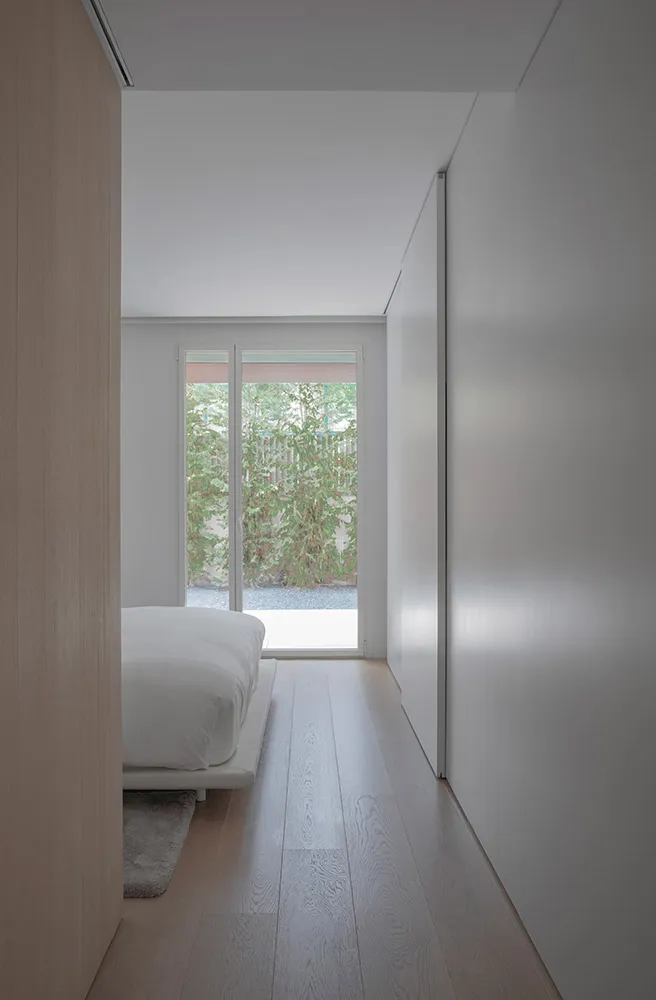 Contemporary bedroom featuring a white bed with minimalist wooden flooring, large windows offering natural light.