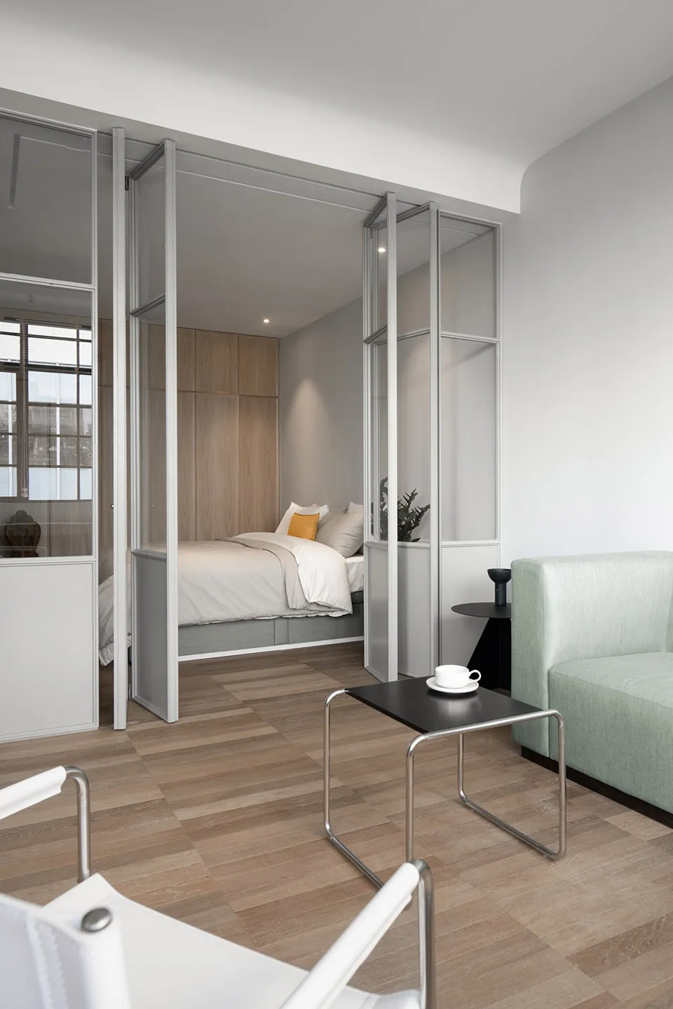 Modern minimalist bedroom with a glass-enclosed sleeping area, wooden flooring, and mint green sofa.