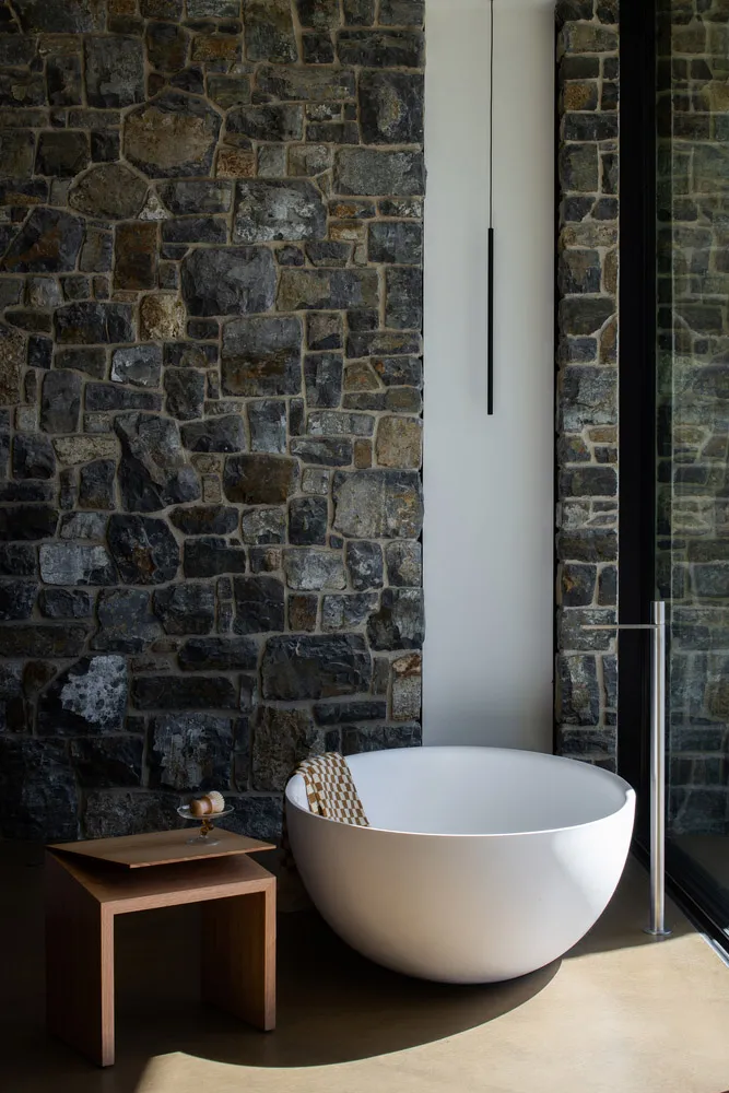 A minimalist bathroom with a stone wall, white bathtub, wooden stool, and glass door.