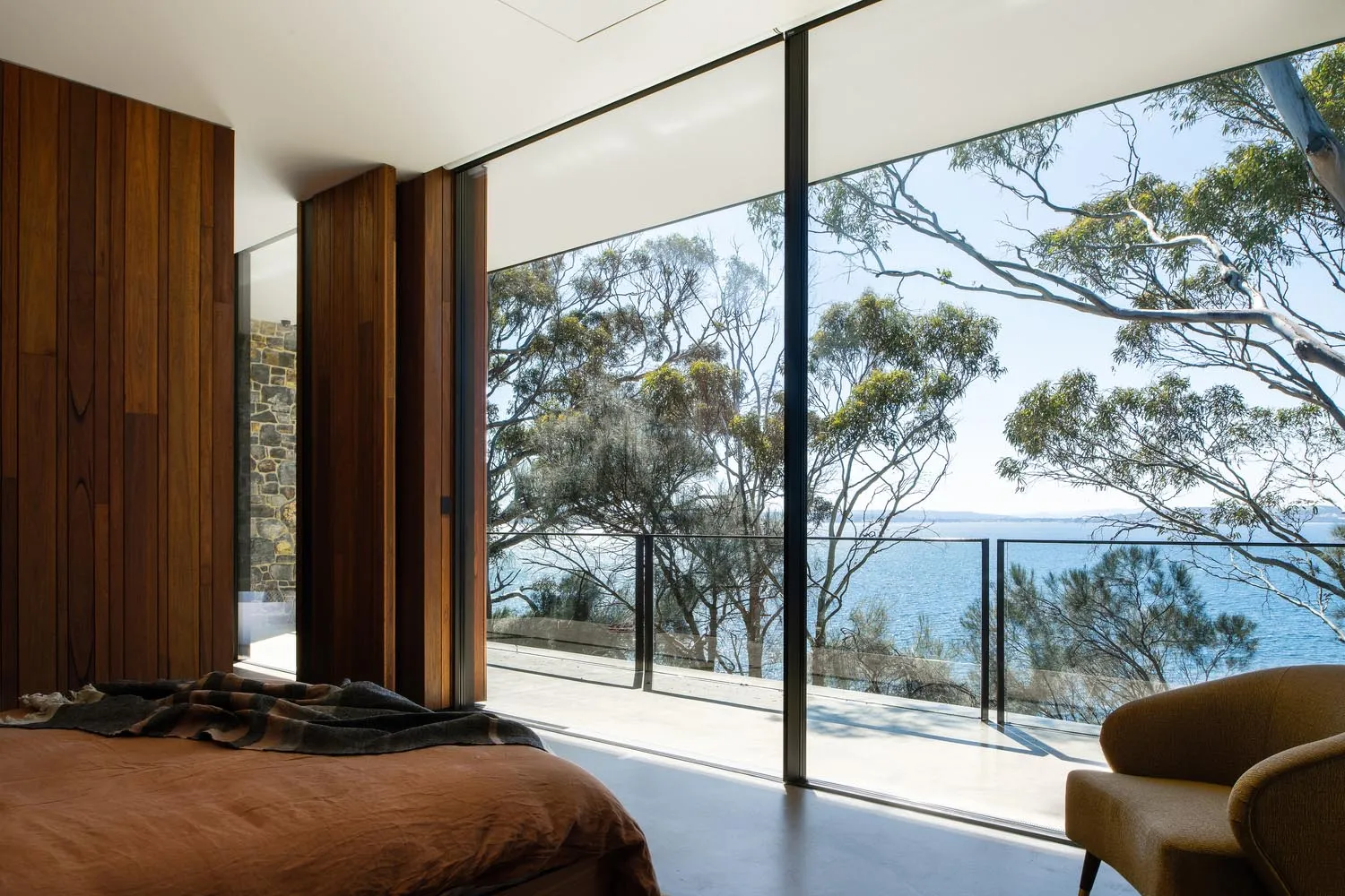 A modern bedroom featuring wood walls, a glass sliding door overlooking the sea, and a concrete floor.