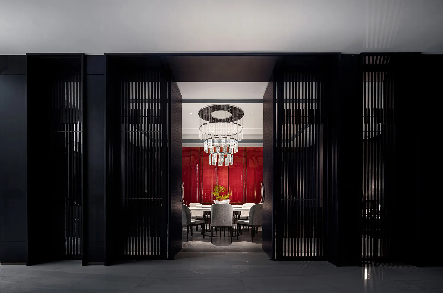 Modern dining room featuring a black glass table, black metal bar, red laminate backdrop, and opulent chandelier hanging from