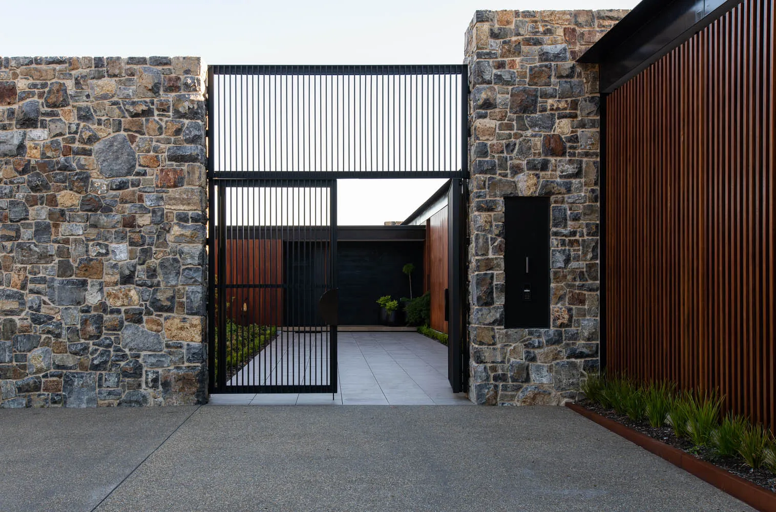 A modern house with stone and concrete exterior features a large gate and a wooden slatted wall.