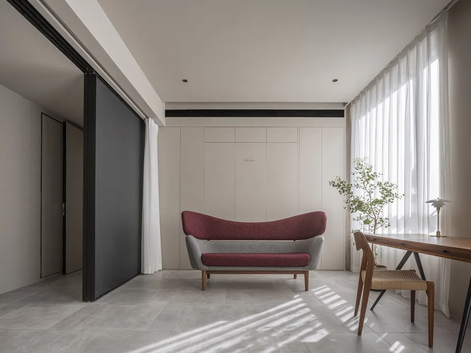 A minimalist living room with a modern sofa, wooden desk, and a plant by the window. The room features neutral-toned walls an