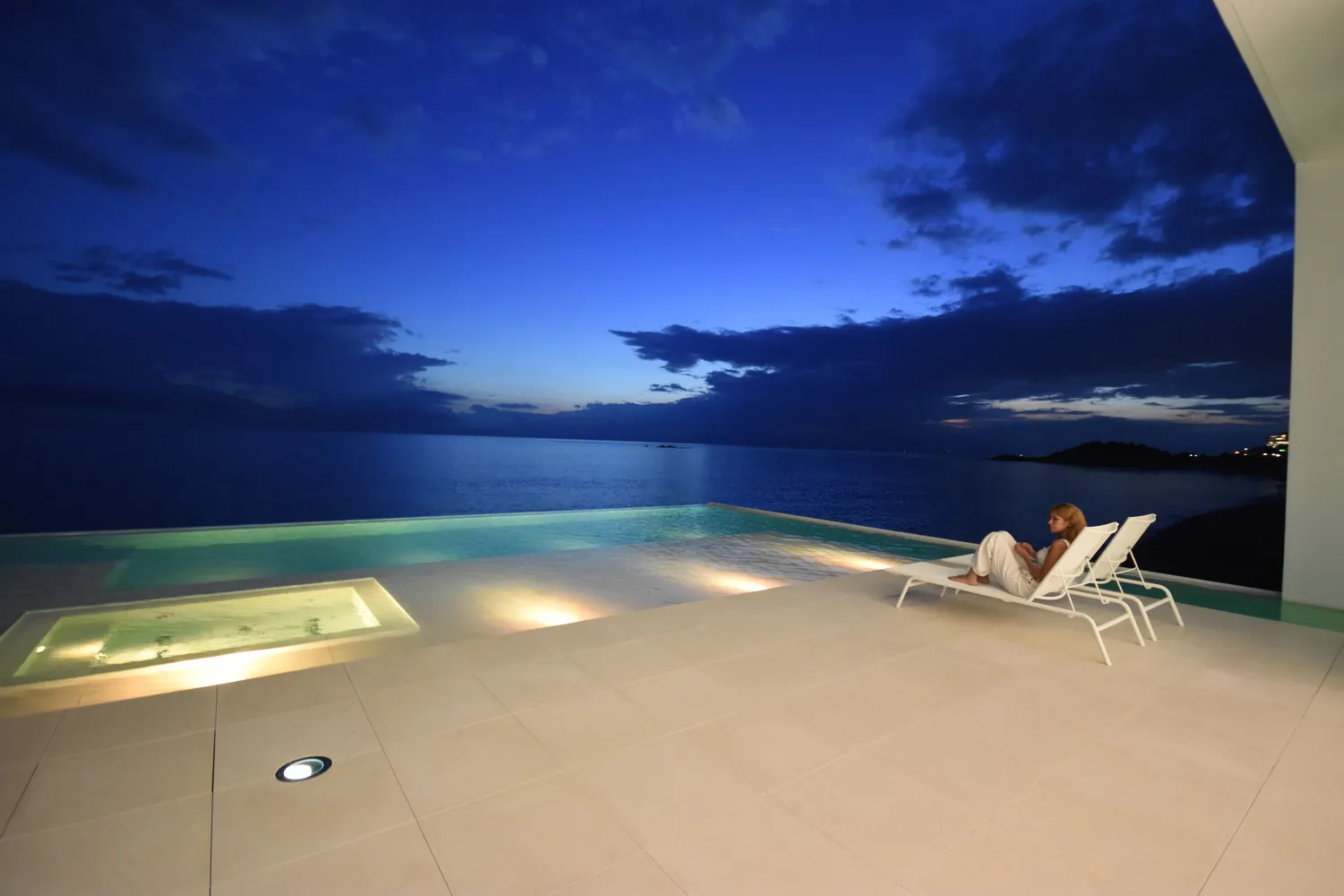 A minimalist view of a waterfront pool at dusk with a person sitting on a lounge chair.