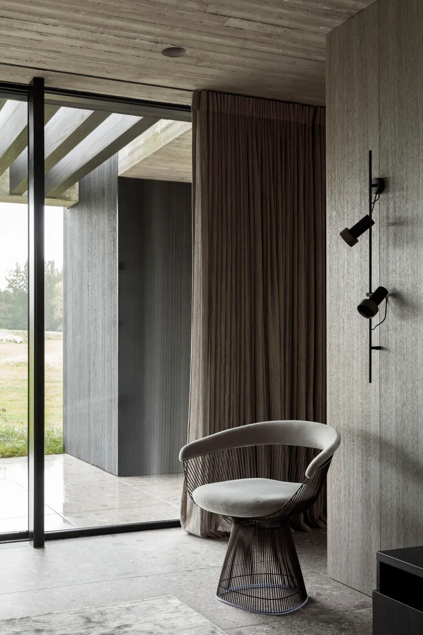 A minimalist interior space featuring a concrete wall, a chair with a wireframe base, and a window with a curtain.