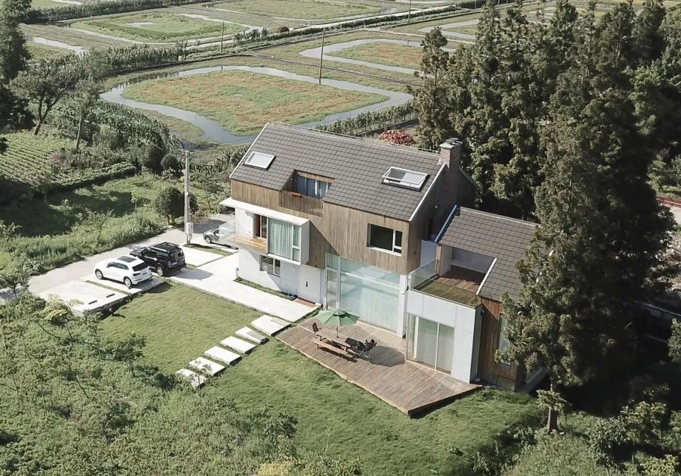 Modern residential villa with wooden frame exterior, surrounded by greenery and a pathway leading to the front door.