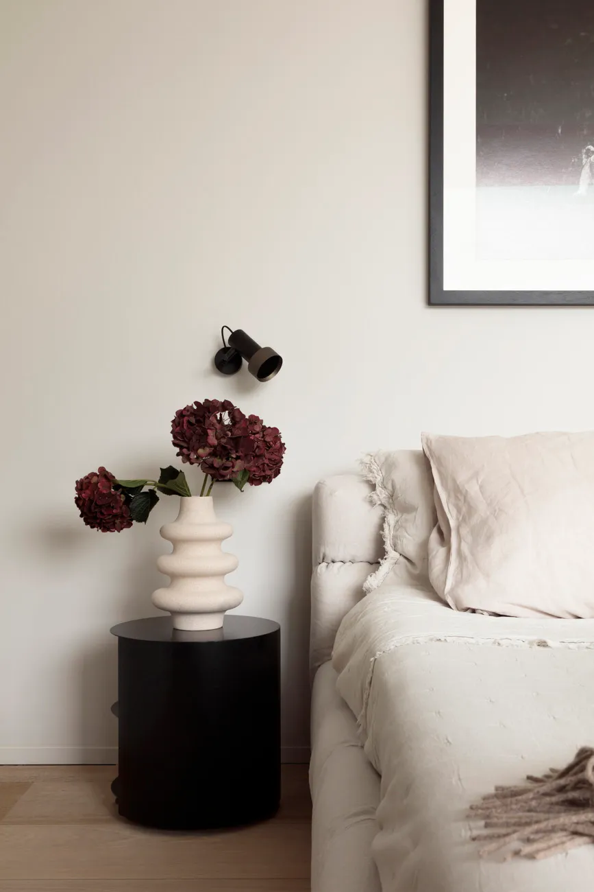 A minimalist bedroom with a white bed featuring textured cushions, situated on a wooden floor with a vase of red flowers on a