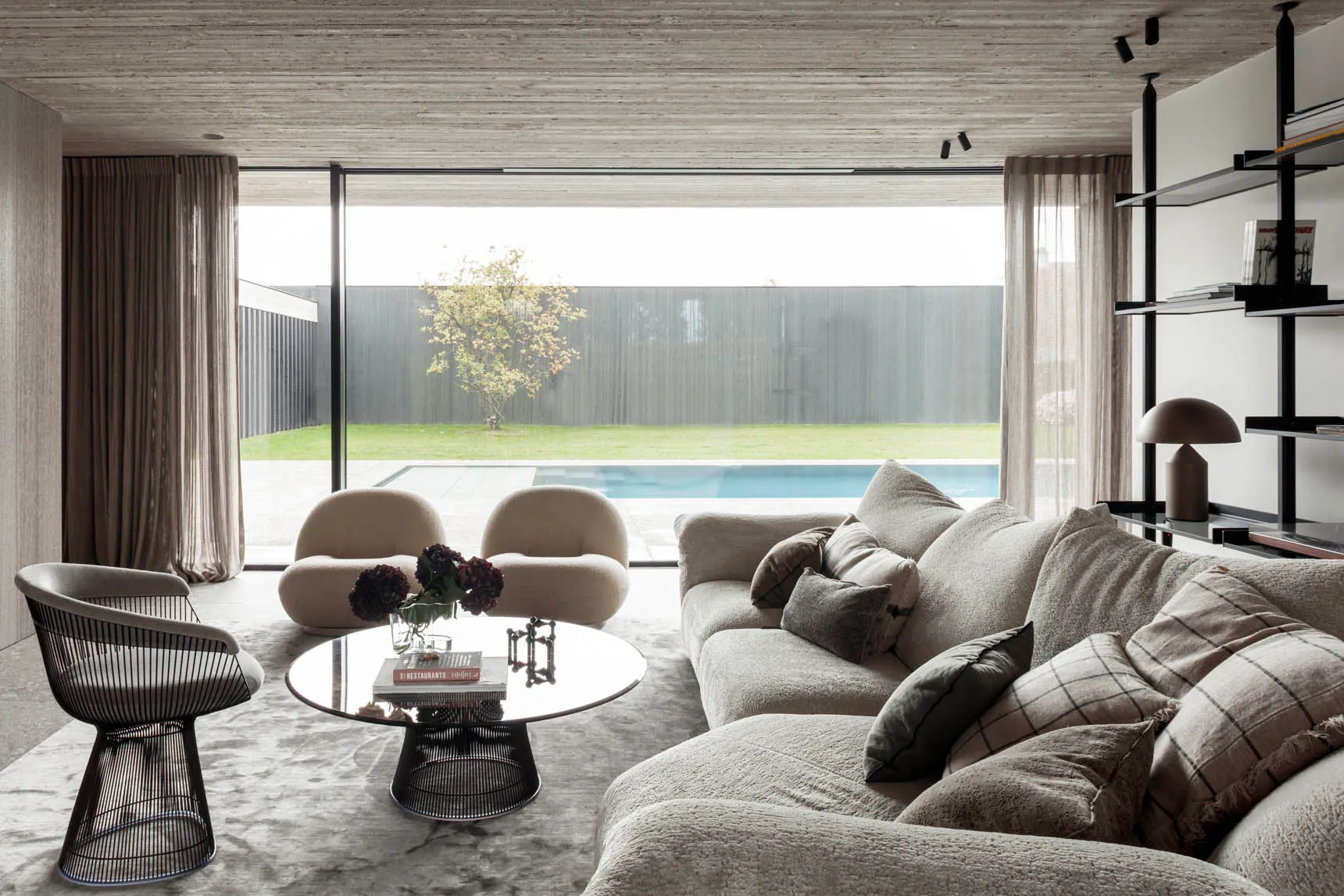 Modern living room with fabric furniture, minimal decor, and large glass windows overlooking a pool.