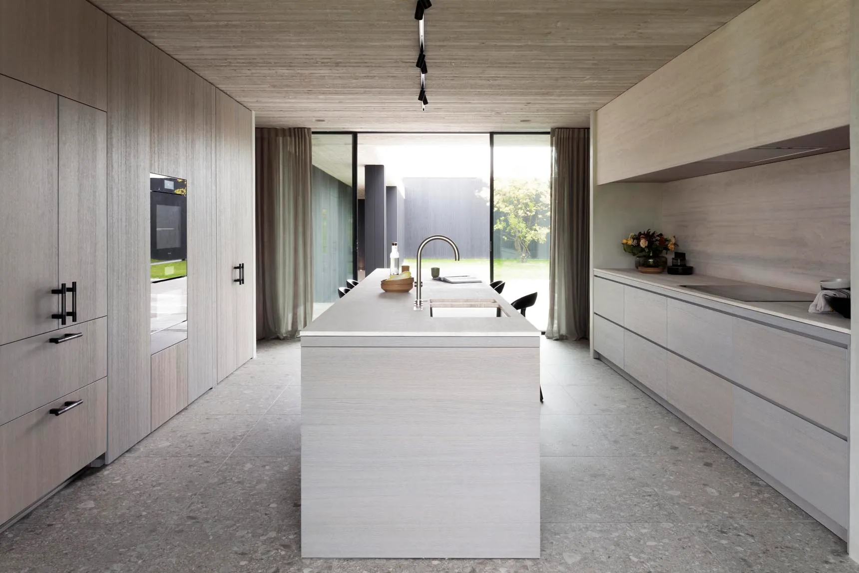 Modern, minimalist kitchen with concrete countertops and floor, featuring an island with a sink and a view of the outdoors th