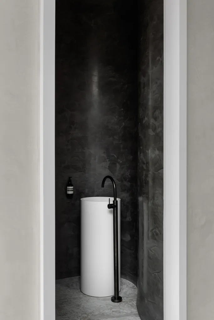 A minimalist bathroom featuring a sleek, cylindrical white sink against a dark, textured concrete wall and floor.