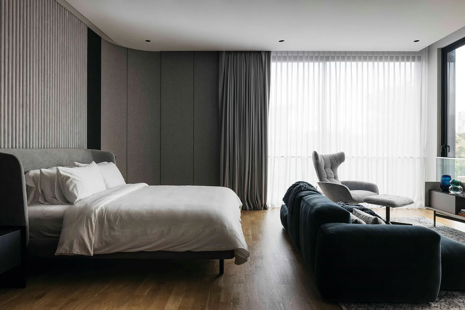 A spacious modern minimalist bedroom featuring textured gray walls, wooden flooring, and a dark sofa with a blanket.