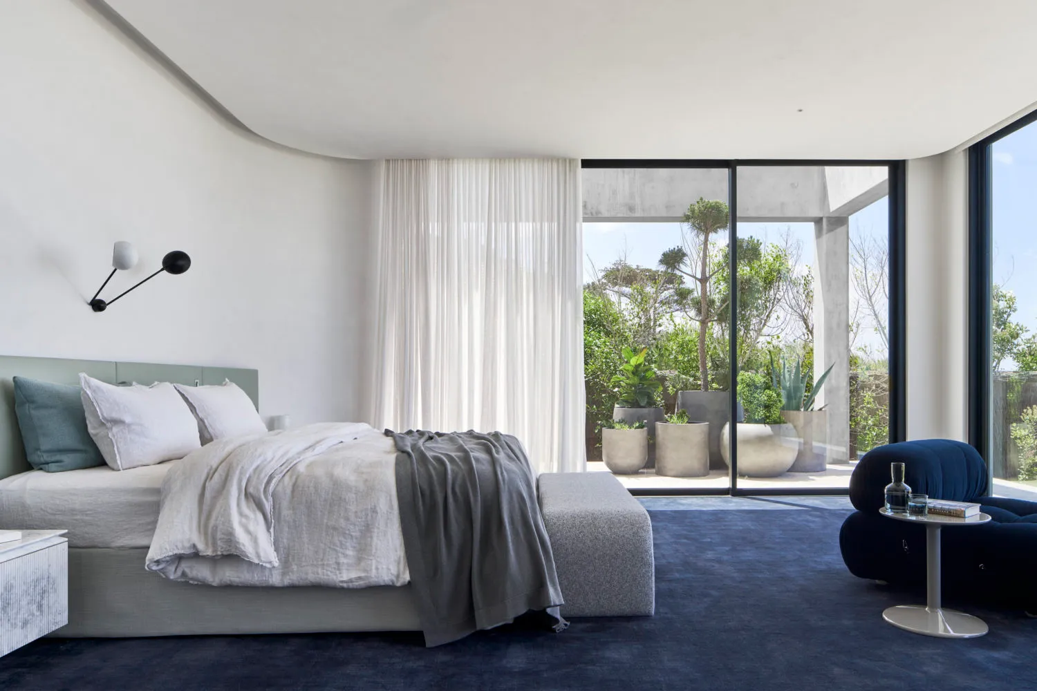 A minimalist bedroom featuring fabric bedding, glass and concrete surfaces, and a large window with a view of greenery outsid