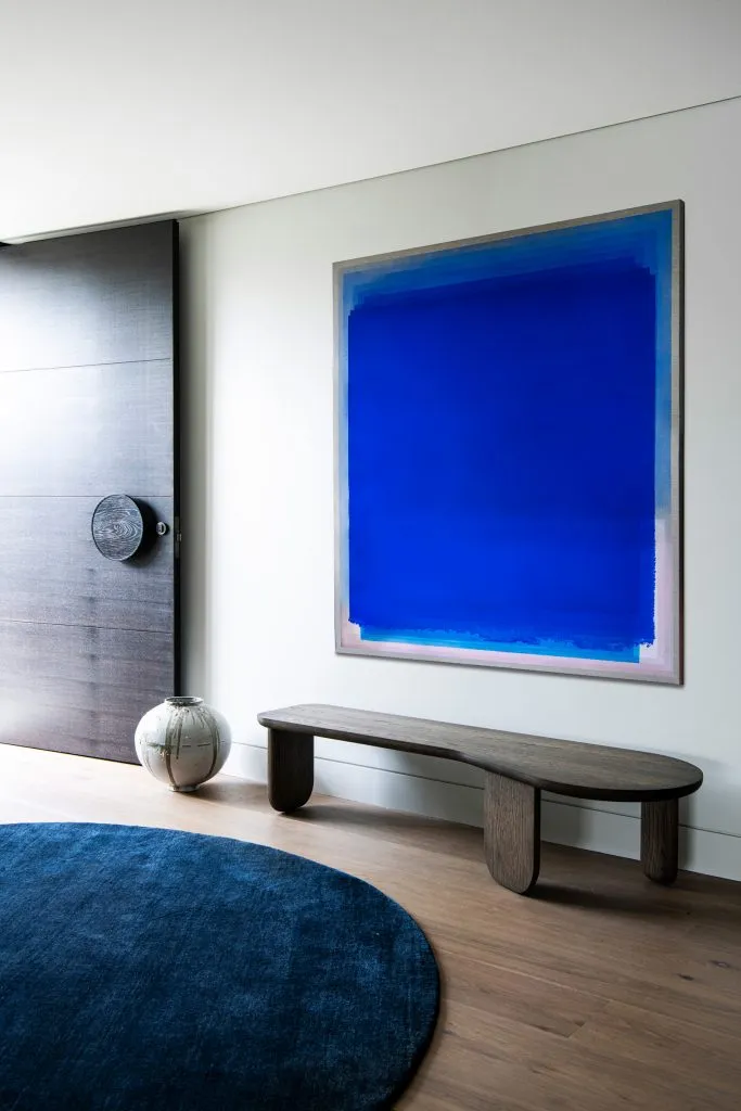 An entryway featuring a blue circular rug, a wooden bench, a blue and pink painting, and a modern wooden door.