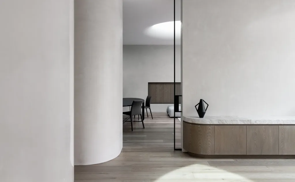 A minimalist dining room with wooden flooring, a wooden table, black chairs, and concrete walls.