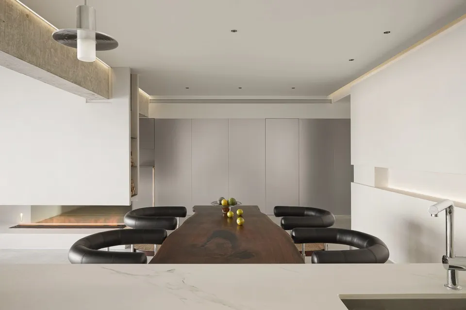 A minimalist dining room featuring a long wooden table with black chairs, surrounded by white walls and a fireplace.