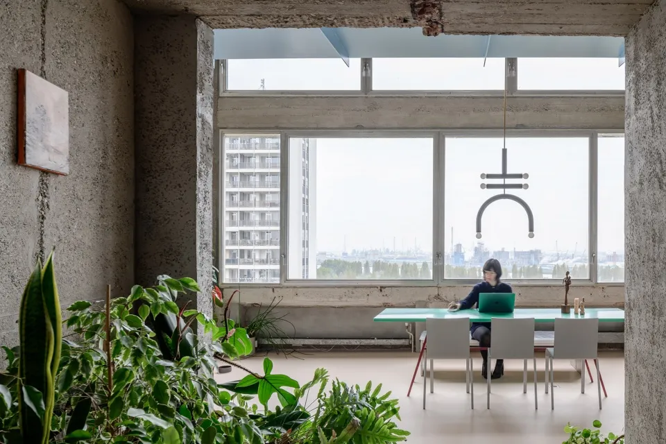 A modern, concrete workspace area with a person working on a laptop, surrounded by large windows overlooking a cityscape.