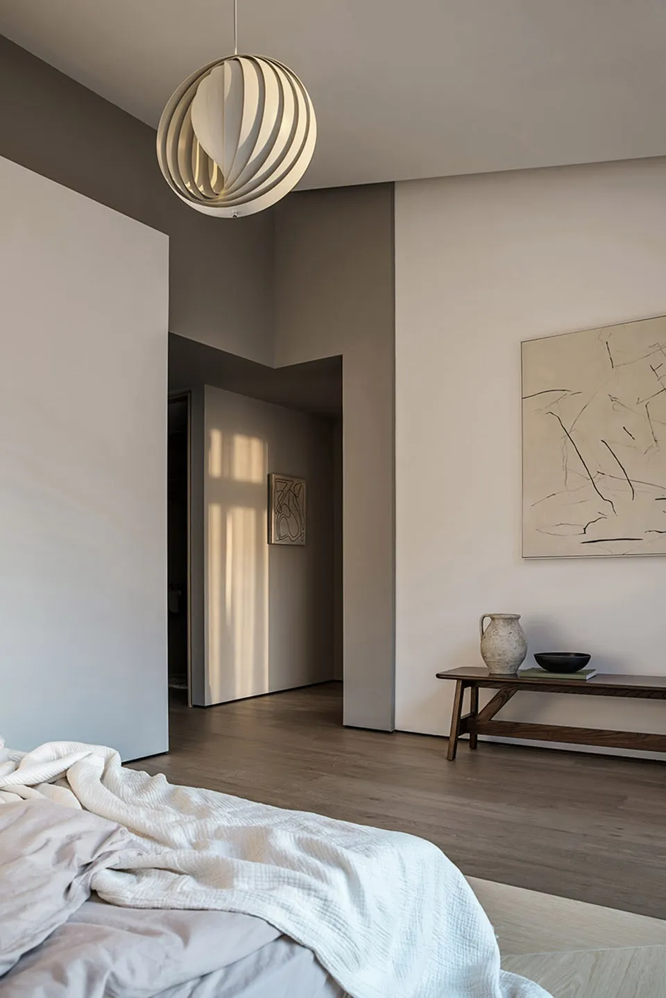 A minimalist bedroom featuring wooden furniture and a cotton blanket, with a modern light fixture and abstract artwork on the