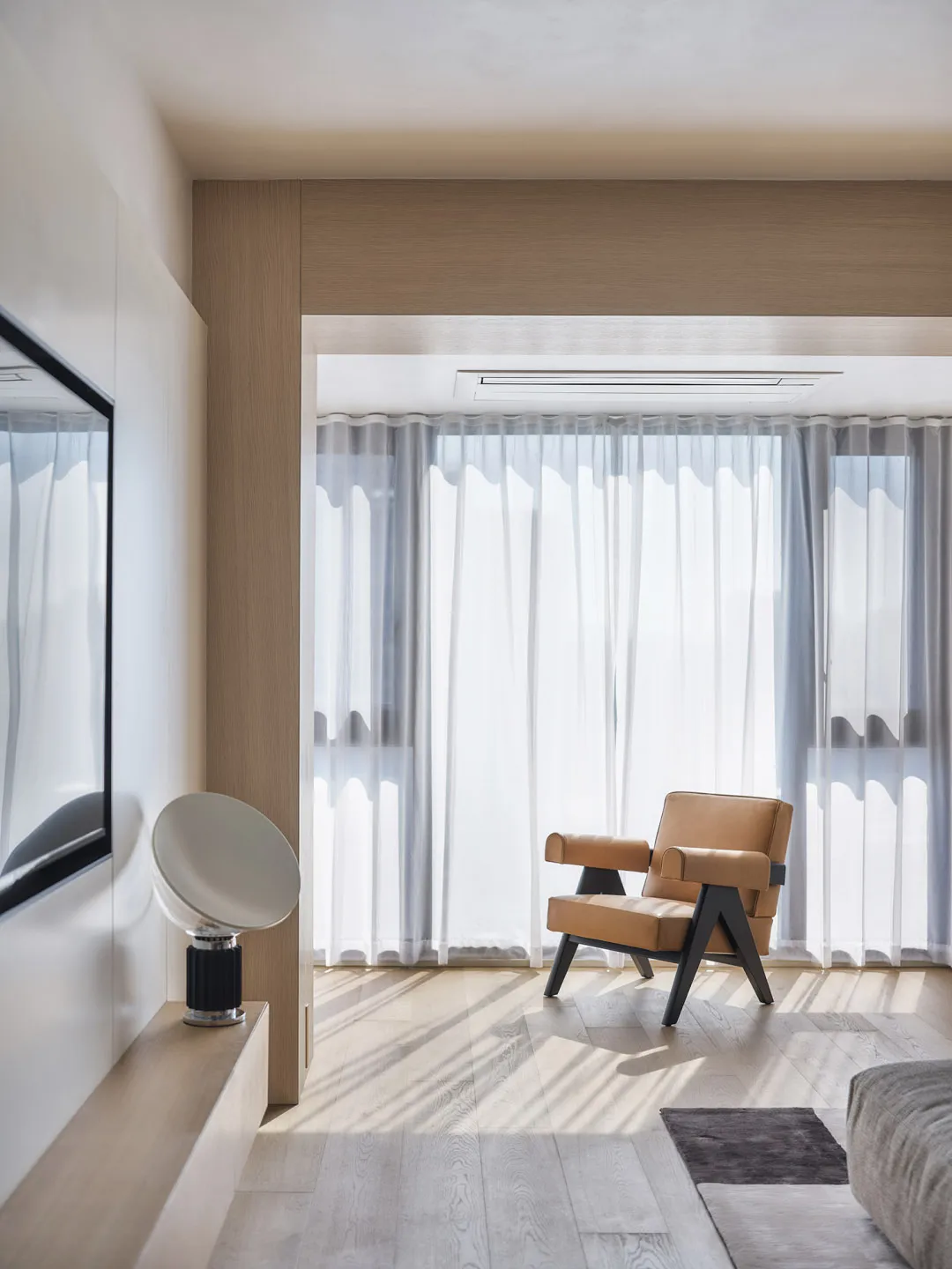 A minimalist living room with a wooden floor, a brown leather armchair, and sheer curtains allowing natural light to filter i