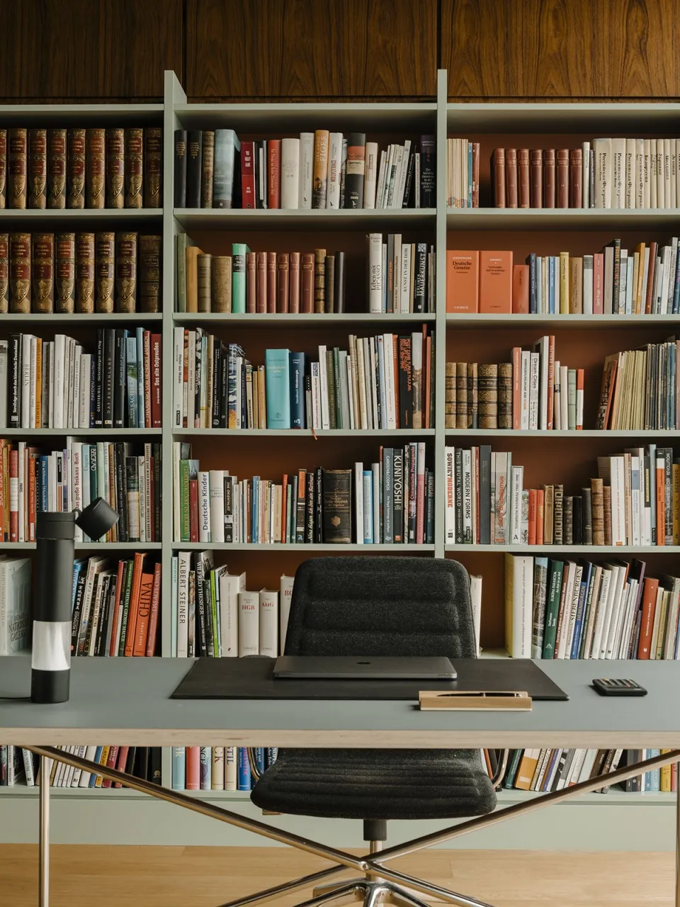 A home office with wooden shelves filled with books and a black desk with a laptop, chair, and lamp.