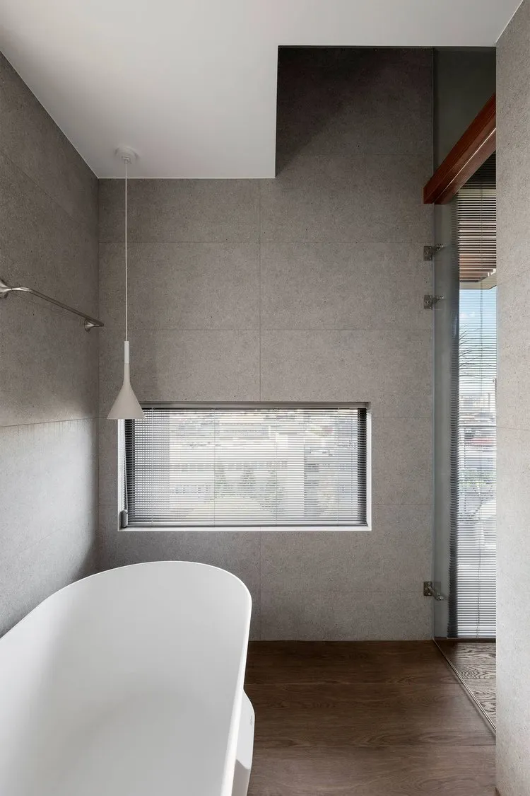 A modern bathroom with a concrete wall, a window with blinds, and a white bathtub.