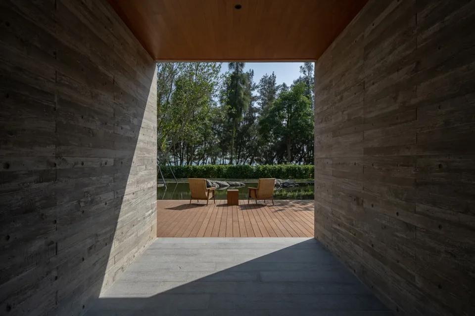 A minimalist landscape featuring wooden deck and stone wall, with a view of trees and water beyond.