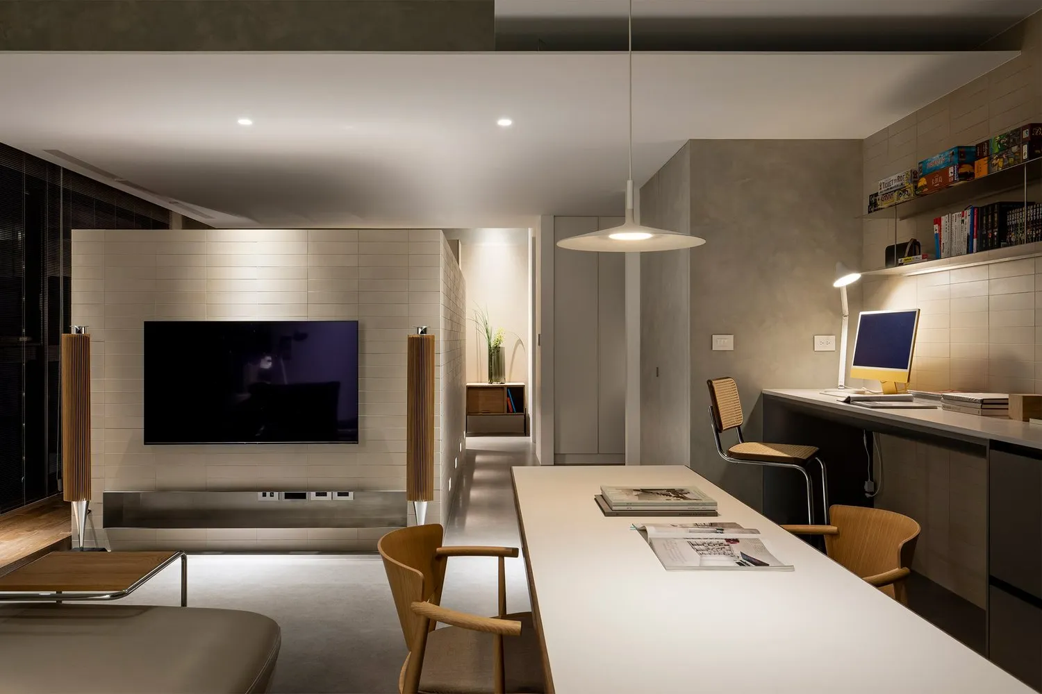 A minimalist living room with a concrete surface, featuring a wall-mounted TV, a long table with a computer setup, and a comf