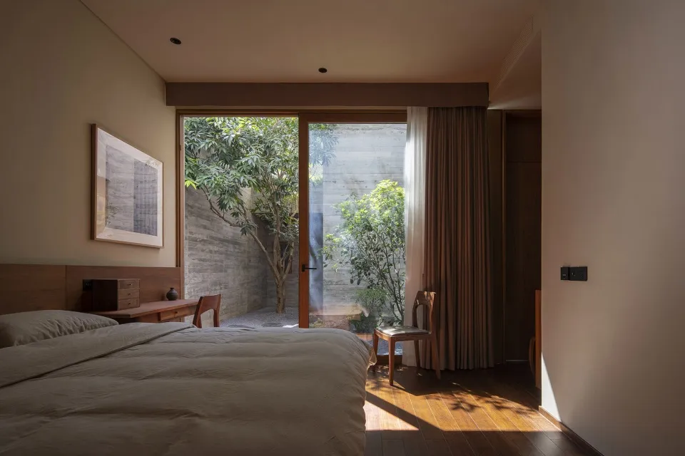 A serene bedroom with a wooden floor, a bed with a wooden headboard, and a large window or sliding glass door leading to a co