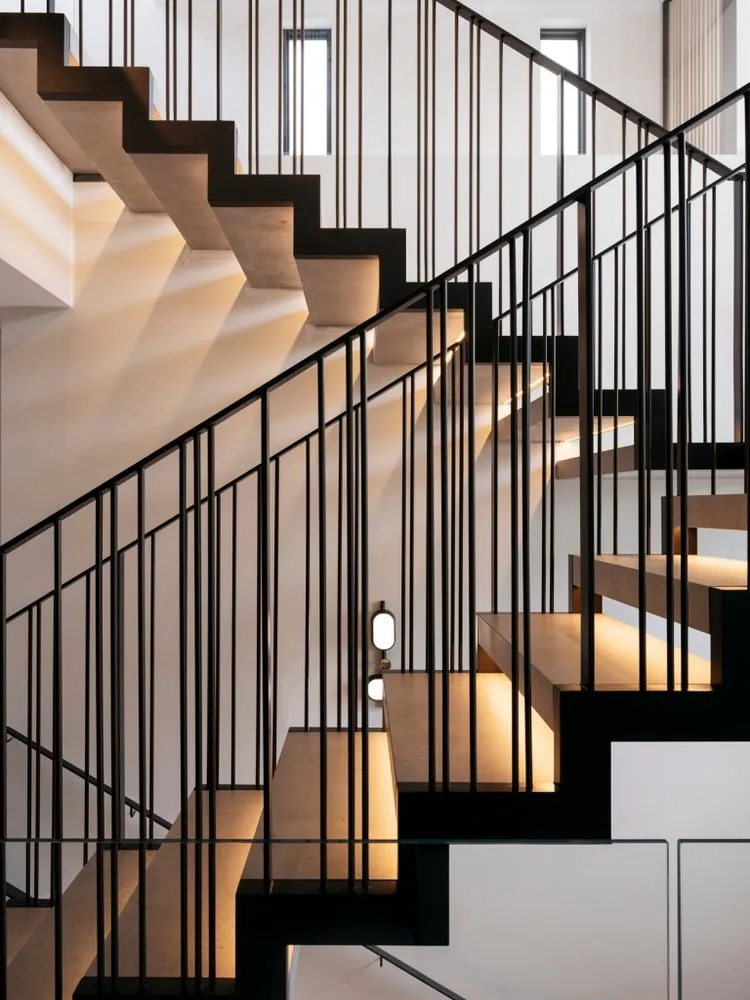 A modern industrial staircase made of concrete with black metal railings, illuminated by soft lighting against a white wall b