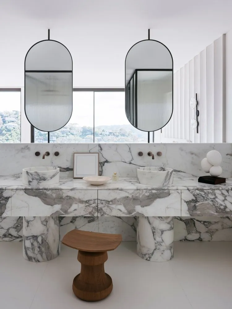 Modern bathroom with marble countertops and sinks, two oval mirrors, and a view of the city.