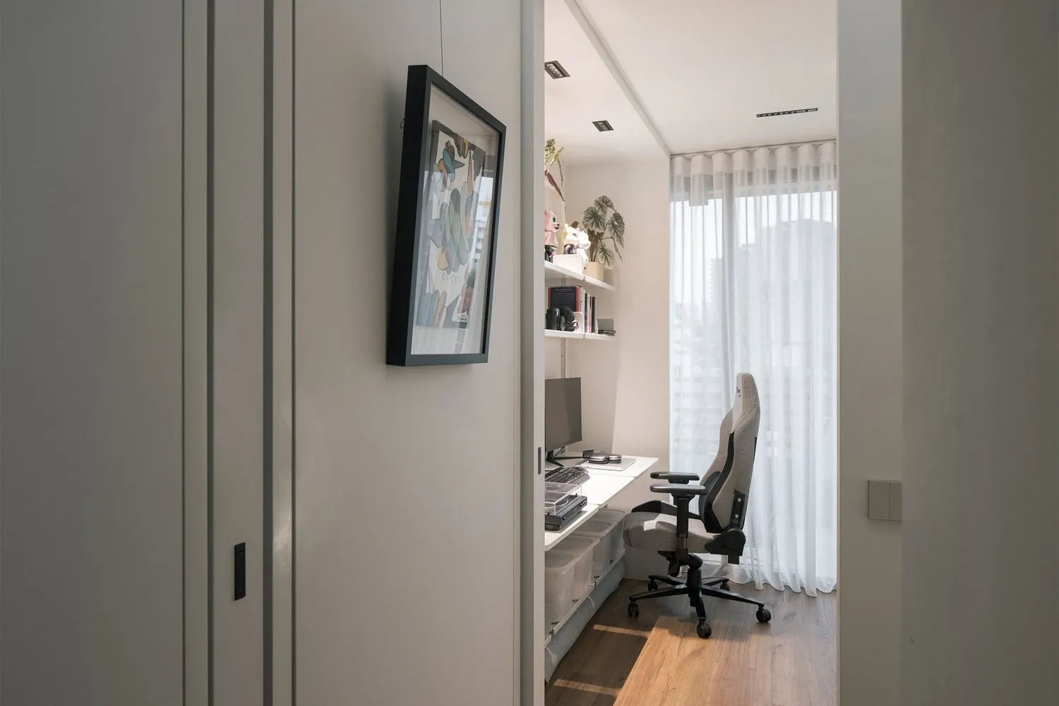 A minimalist workspace with a white desk, grey chair, and a framed artwork hanging on the wall with a white curtain in the ba