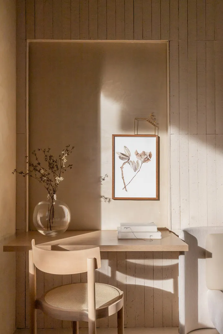 A minimalist workspace featuring a wooden desk, chair, vase with dried flowers, and a framed botanical print on the wall, wit