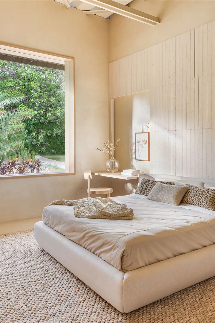 A minimalist bedroom featuring neutral colors with a large window overlooking greenery and a small workspace area.