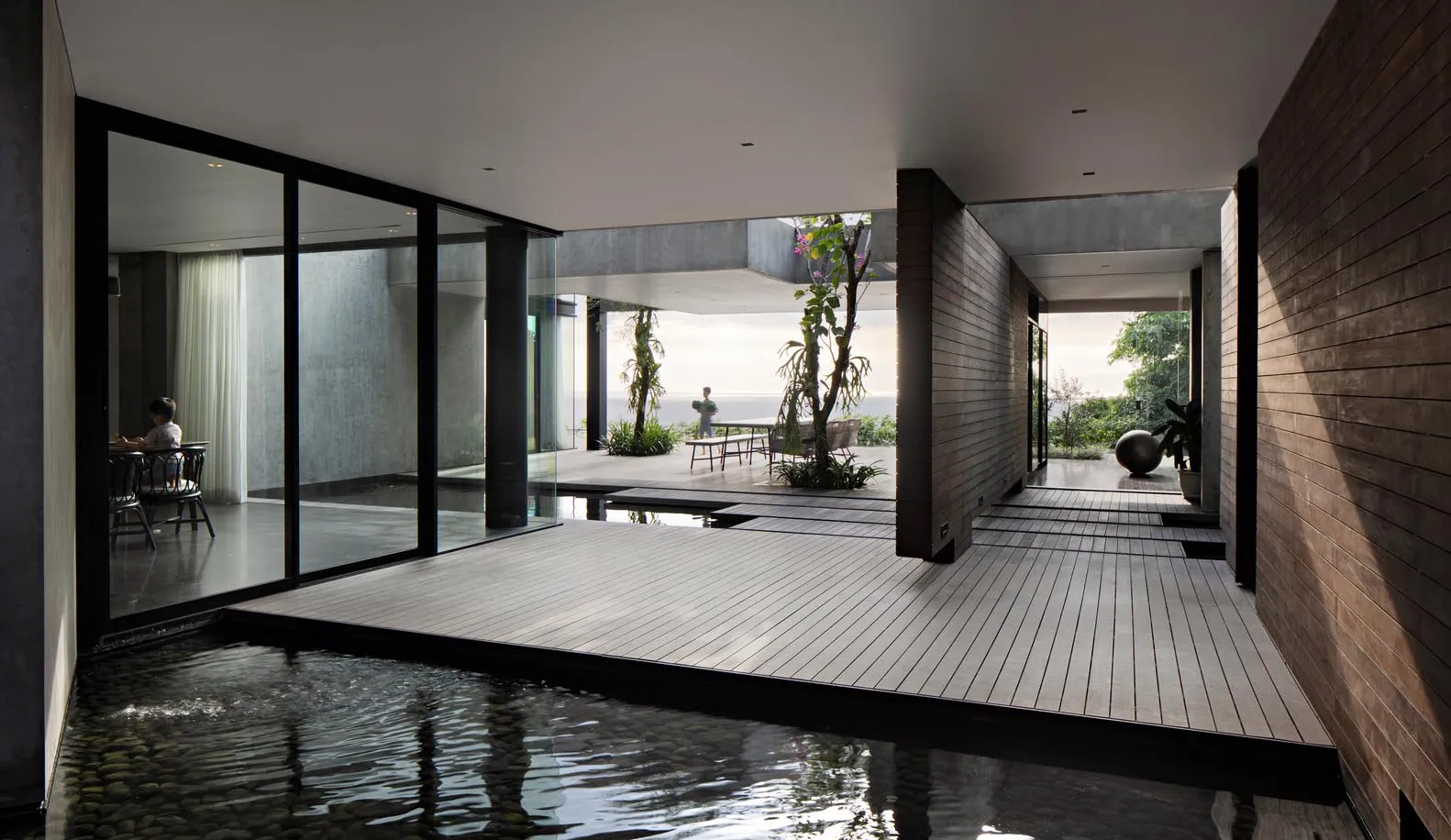 Modern residential living room with glass walls, wood floor, and concrete columns overlooking an outdoor space.