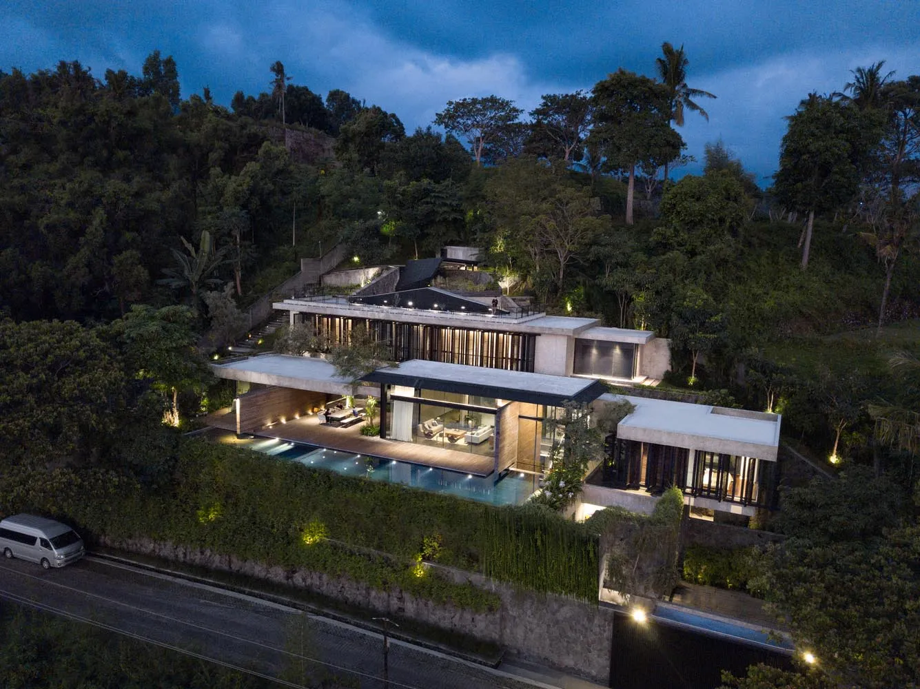 A modern residential lounge featuring a concrete and glass structure with a swimming pool at dusk.