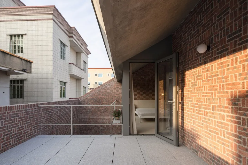 A modern residential balcony with a brick wall on the left and concrete flooring. The interior reveals a minimalist bedroom s