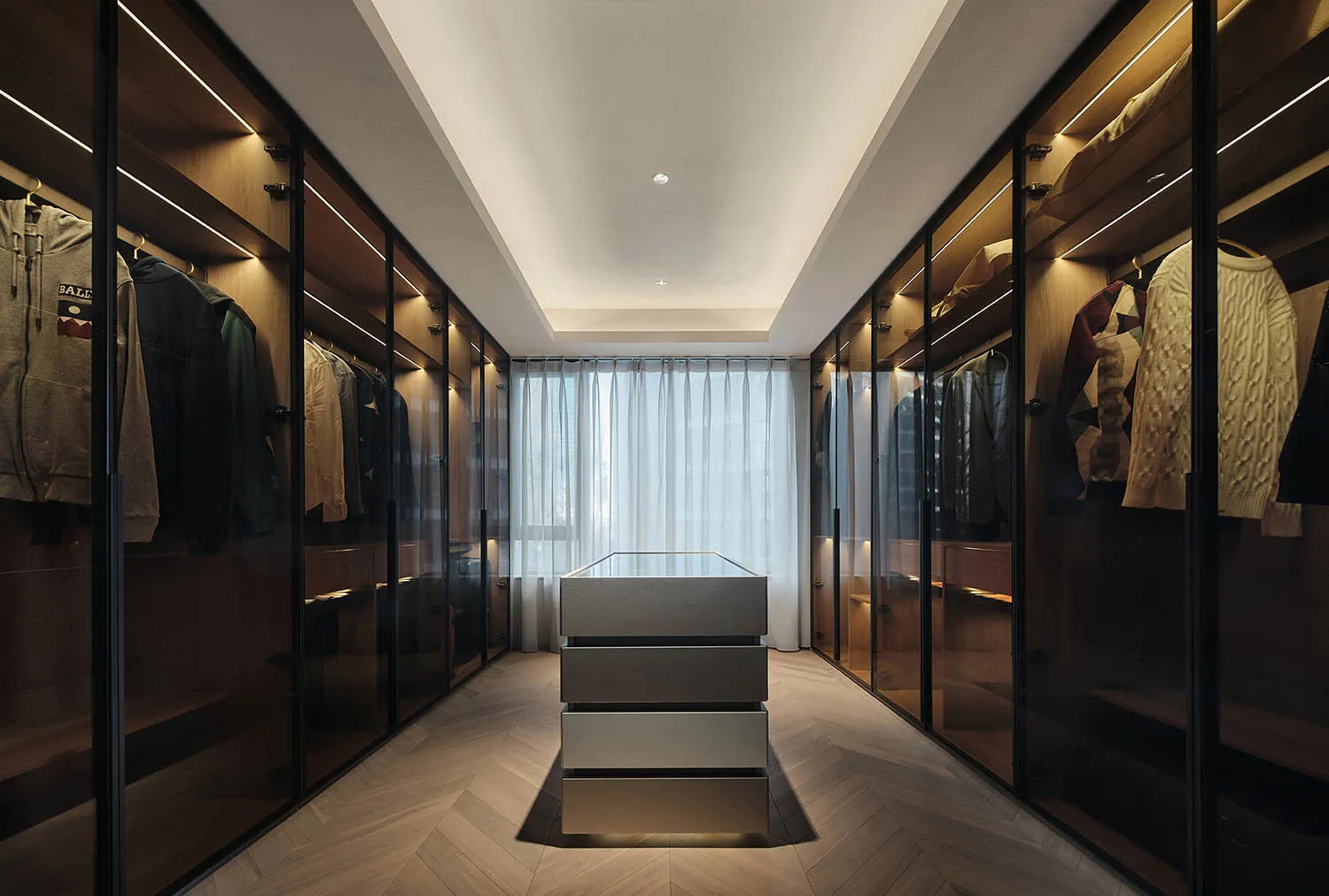 Modern walk-in closet with glass doors, white drawers, and wooden flooring.