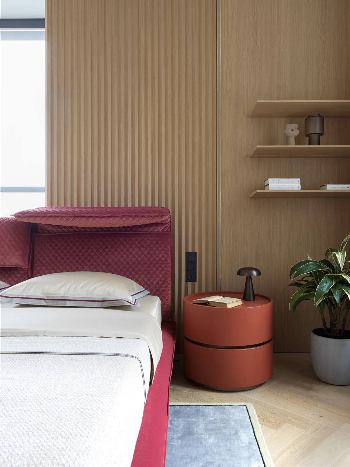 A minimalistic bedroom with a red velvet bed frame, wooden wall, and a circular bedside table with a plant.