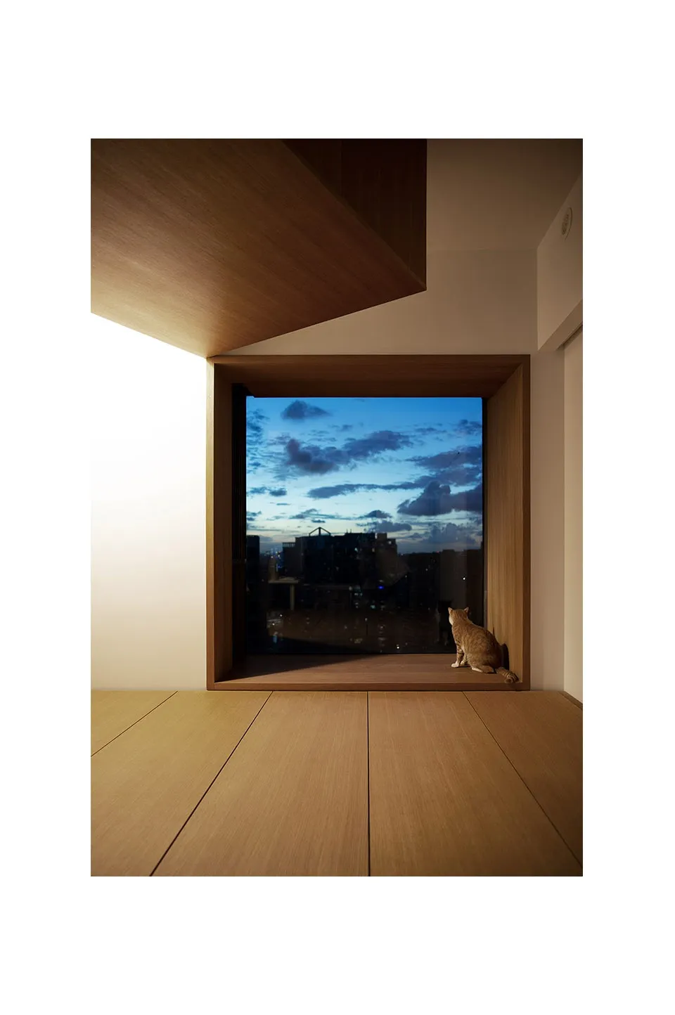 A modern living space featuring a wooden floor and a large window overlooking a cityscape during sunset.