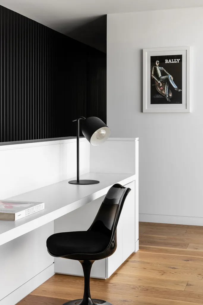 A minimalist home office features a white desk with a black lamp and chair, set against a black and white striped wall and wo