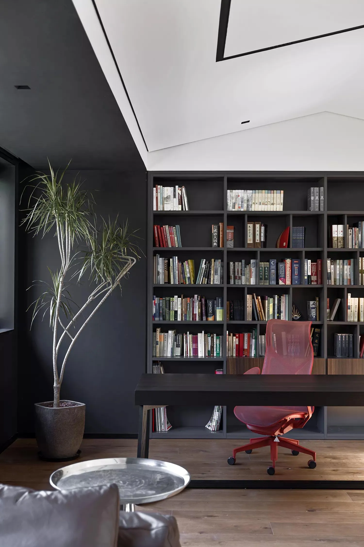 A minimalist home library with a large bookshelf filled with books, a wooden desk, and a red office chair.