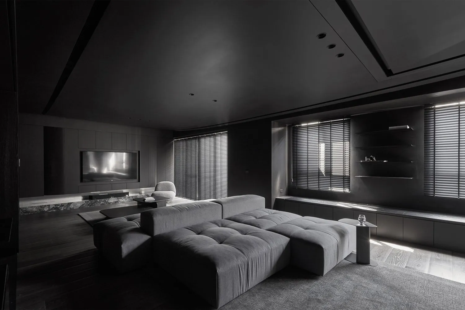 Minimalist living room interior with a upholstered grey couch, recessed lighting, and black window shutters.